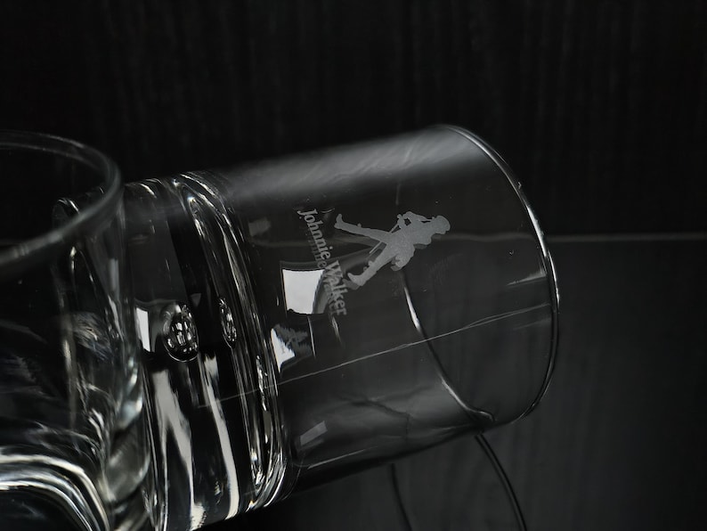 May include: A clear, cylindrical glass with the "Johnnie Walker" logo etched on the side. The logo features a silhouette of a man walking. The glass is partially filled and angled against a dark background.