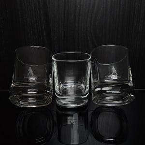 May include: Three clear, lowball whiskey glasses with a distinctive base and a logo of a striding figure. The glasses are arranged on a reflective black surface against a dark background. The central glass is a square shape.