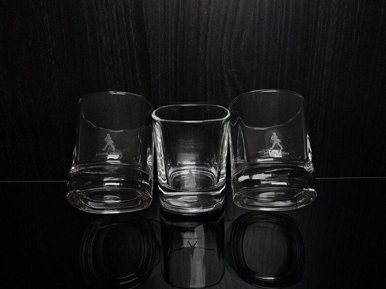 May include: Three clear, lowball whiskey glasses with a distinctive base and a logo of a striding figure. The glasses are arranged on a reflective black surface against a dark background. The central glass is a square shape.