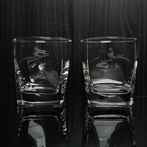 May include: Two clear, square-shaped whiskey glasses with etched "Johnnie Walker" branding and a figure of a man walking. The glasses are reflected on a dark, glossy surface, with a dark wood-grain background.