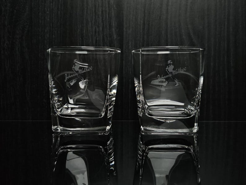 May include: Two clear, square-shaped whiskey glasses with etched "Johnnie Walker" branding and a figure of a man walking. The glasses are reflected on a dark, glossy surface, with a dark wood-grain background.