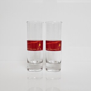 May include: Two clear, tall drinking glasses with a red band featuring a walking figure and the text "Johnnie Walker." The glasses are empty and reflect on the white surface below. The design is a classic for whiskey enthusiasts.