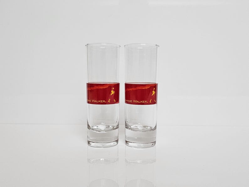 May include: Two clear, tall drinking glasses with a red band featuring a walking figure and the text "Johnnie Walker." The glasses are empty and reflect on the white surface below. The design is a classic for whiskey enthusiasts.
