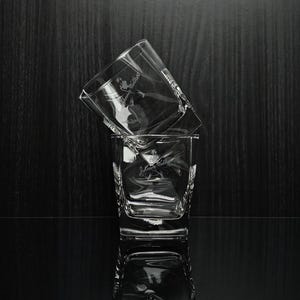 May include: Two clear, square-shaped whiskey glasses stacked on a reflective surface. Each glass has a logo etched on the front. The background is a dark wood grain.