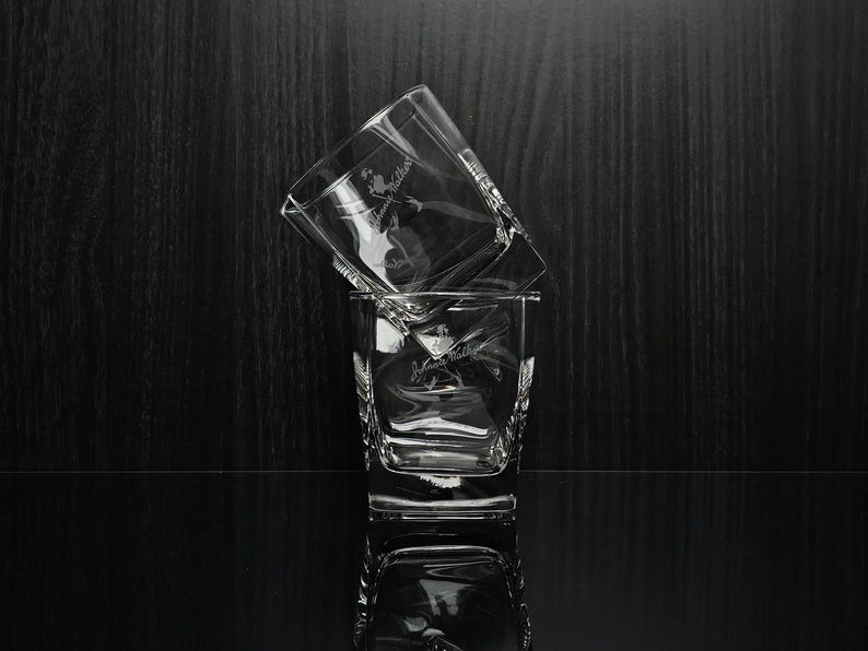 May include: Two clear, square-shaped whiskey glasses stacked on a reflective surface. Each glass has a logo etched on the front. The background is a dark wood grain.