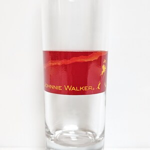 May include: A clear glass tumbler with a red band featuring the "Johnnie Walker" logo in gold. The band has a textured, marbled appearance. The glass is cylindrical and appears to be empty.