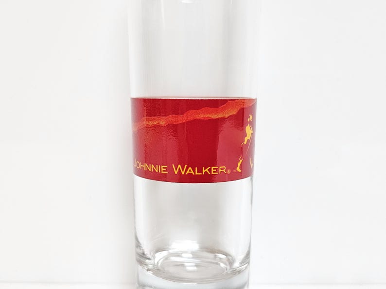 May include: A clear glass tumbler with a red band featuring the "Johnnie Walker" logo in gold. The band has a textured, marbled appearance. The glass is cylindrical and appears to be empty.