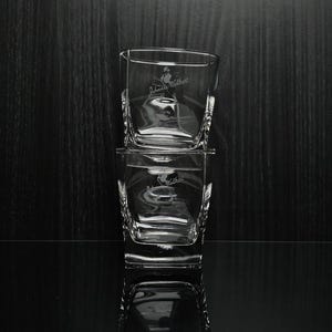 May include: Two clear, square-shaped whiskey glasses stacked on top of each other. Each glass has a logo etched on the front. The glasses are on a reflective black surface with a dark wood background.