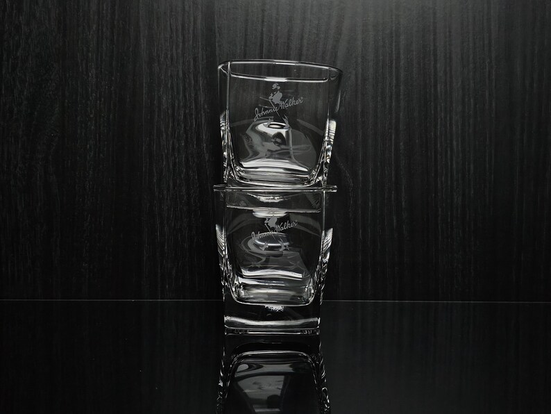 May include: Two clear, square-shaped whiskey glasses stacked on top of each other. Each glass has a logo etched on the front. The glasses are on a reflective black surface with a dark wood background.