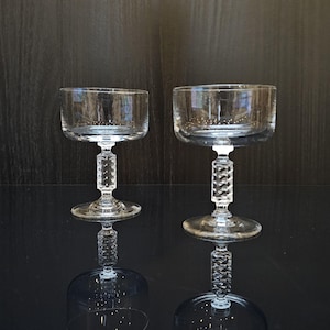 Op de afbeelding: Twee heldere glazen champagne coupes met een getextureerde steel. De glazen staan op een zwart oppervlak.