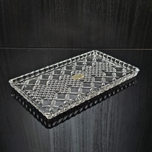 Könnte beinhalten: Ein rechteckiges Tablett aus klarem Glas mit einem detaillierten, gepressten Rauten- und Kreuzschraffurmuster auf der Oberfläche. Die Ränder des Tabletts sind gewellt und erhöht, und in der Mitte befindet sich ein kleiner goldfarbener Aufkleber.