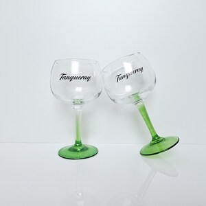 May include: Two clear gin glasses with green stems and bases, each bearing the word "Tanqueray" in black script. The glasses are positioned against a white background, one upright and the other tilted.