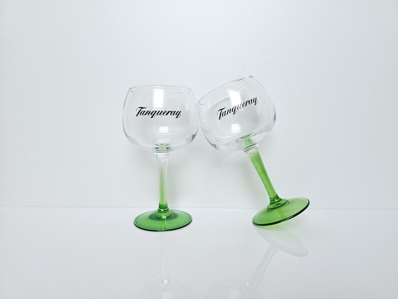 May include: Two clear gin glasses with green stems and bases, each bearing the word "Tanqueray" in black script. The glasses are positioned against a white background, one upright and the other tilted.