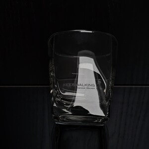 May include: A clear, square-shaped whiskey glass with the text "KEEP WALKING" and "JOHNNIE WALKER" etched on the side. The glass has a textured design and is set against a dark background.