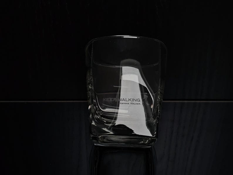 May include: A clear, square-shaped whiskey glass with the text "KEEP WALKING" and "JOHNNIE WALKER" etched on the side. The glass has a textured design and is set against a dark background.