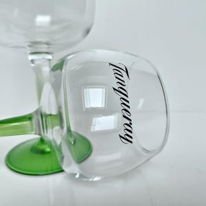 May include: Two clear, stemmed glasses with green bases. One glass is upright, the other is tilted, displaying the word "Tanqueray" in black script on the side. The glasses are on a white surface.