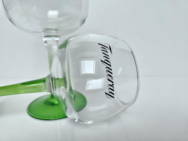 May include: Two clear, stemmed glasses with green bases. One glass is upright, the other is tilted, displaying the word "Tanqueray" in black script on the side. The glasses are on a white surface.