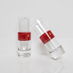 May include: Two clear shot glasses with a red band featuring the text "Johnnie Walker" and a logo. One glass is upright, the other is tilted. The glasses are partially filled with a clear liquid.