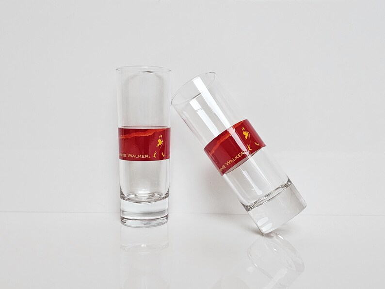 May include: Two clear shot glasses with a red band featuring the text "Johnnie Walker" and a logo. One glass is upright, the other is tilted. The glasses are partially filled with a clear liquid.