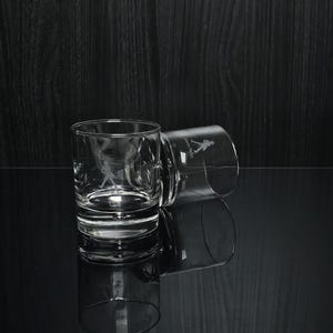 May include: Two clear, lowball glasses with etched images of a golfer in action. One glass is upright, the other tilted, both reflecting on a glossy black surface. The glasses are designed for spirits.