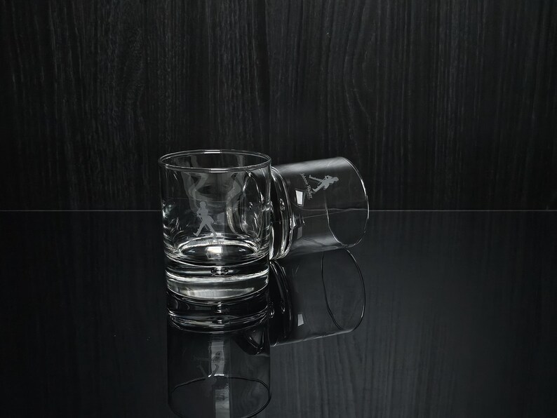 May include: Two clear, lowball glasses with etched images of a golfer in action. One glass is upright, the other tilted, both reflecting on a glossy black surface. The glasses are designed for spirits.