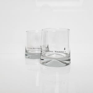 Peut inclure: Deux verres &agrave; whisky transparents, de forme cylindrique, portant le logo "Johnnie Walker" et un embl&egrave;me d'homme en marche. Les verres ont une base &eacute;paisse et refl&egrave;tent la lumi&egrave;re. Fond blanc.