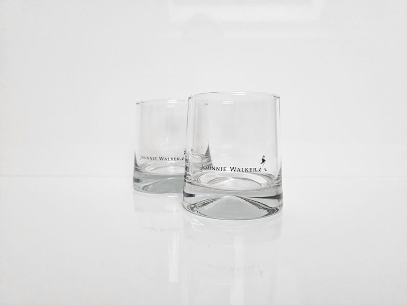 Peut inclure: Deux verres &agrave; whisky transparents, de forme cylindrique, portant le logo "Johnnie Walker" et un embl&egrave;me d'homme en marche. Les verres ont une base &eacute;paisse et refl&egrave;tent la lumi&egrave;re. Fond blanc.