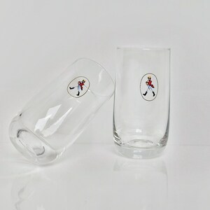 May include: Two clear, cylindrical drinking glasses. Each glass has a small, oval sticker with a cartoon figure in a red coat. One glass is upright, the other is tilted to the side.