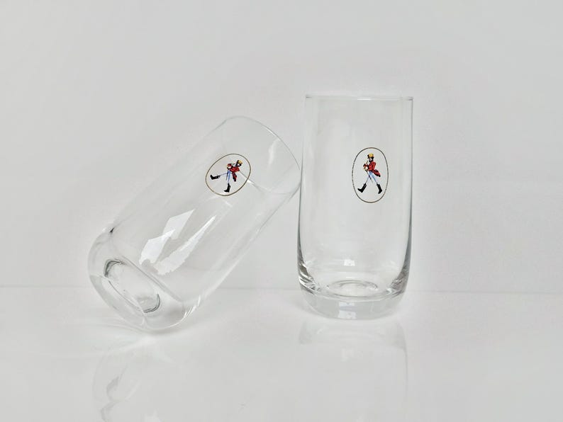 May include: Two clear, cylindrical drinking glasses. Each glass has a small, oval sticker with a cartoon figure in a red coat. One glass is upright, the other is tilted to the side.