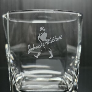 May include: Clear square-shaped glass with the etched Johnnie Walker logo. The logo features a silhouette of a man walking with a cane. The glass has a textured design on the sides.