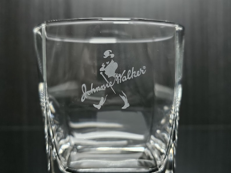 May include: Clear square-shaped glass with the etched Johnnie Walker logo. The logo features a silhouette of a man walking with a cane. The glass has a textured design on the sides.