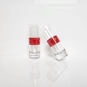 May include: Two clear shot glasses with a red band featuring gold text and a logo. One glass is upright, the other is tilted. The glasses are partially filled with a clear liquid, and sit on a white surface.