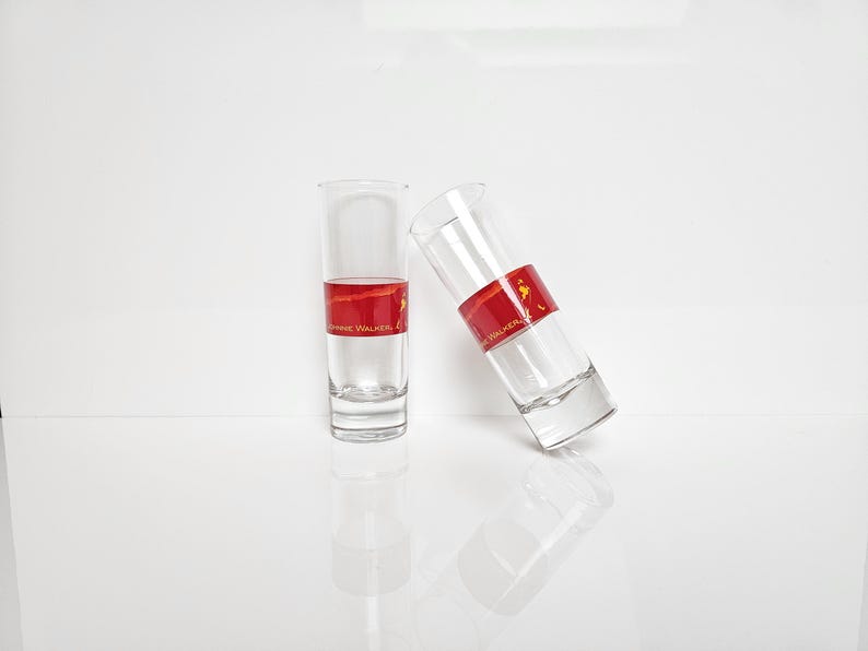 May include: Two clear shot glasses with a red band featuring gold text and a logo. One glass is upright, the other is tilted. The glasses are partially filled with a clear liquid, and sit on a white surface.