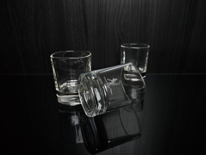 May include: Three clear, square-shaped shot glasses are arranged on a reflective black surface. One glass is lying on its side, displaying an etched design. The other two glasses are upright, with the background a dark wood.