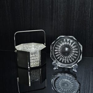 Op de afbeelding: Een set doorzichtige glazen onderzetters met een zonnestraalpatroon, geplaatst in een gepolijste zilverkleurige metalen houder met een zwenkbare handgreep. Eén onderzetter staat rechtop op een doorzichtige standaard, de rest zit in de houder.