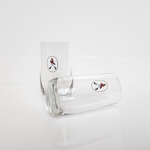 May include: Two clear, rectangular highball glasses with a small, oval logo of a figure in a red coat and black pants. One glass is upright, the other is lying on its side. The glasses are on a white surface.