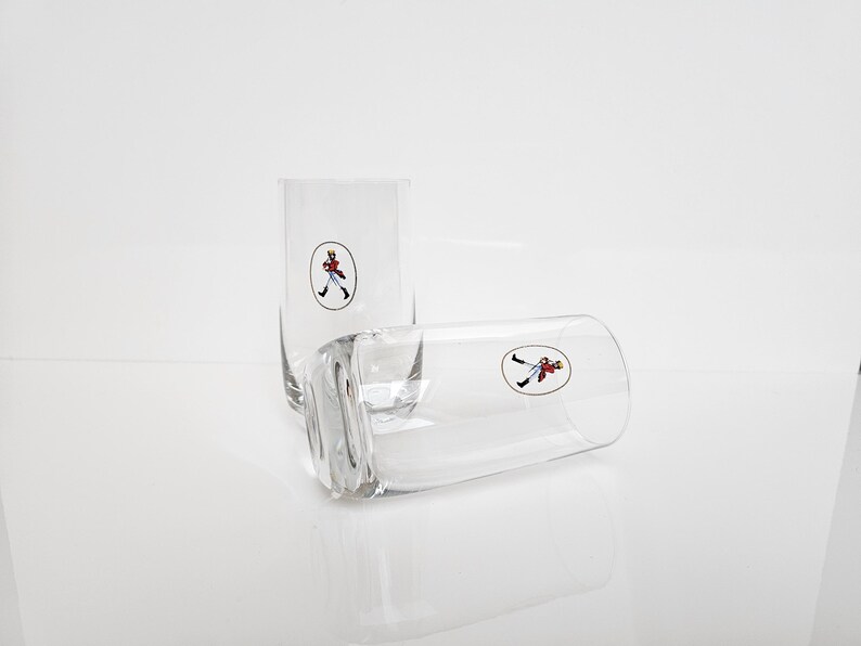 May include: Two clear, rectangular highball glasses with a small, oval logo of a figure in a red coat and black pants. One glass is upright, the other is lying on its side. The glasses are on a white surface.