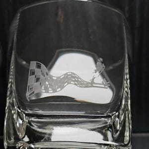 May include: Clear square glass with an etched design. The design features a checkered flag waving in the wind, with a figure walking on a diagonal line. The glass is likely a whiskey tumbler.