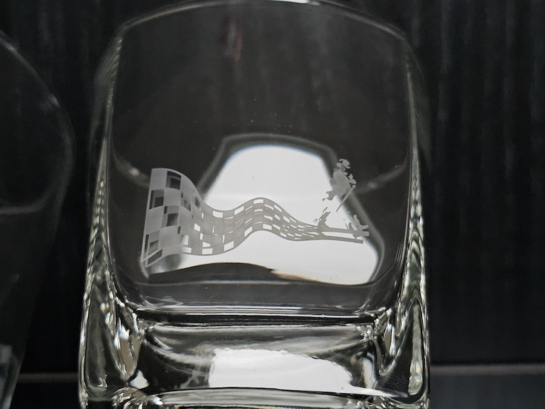 May include: Clear square glass with an etched design. The design features a checkered flag waving in the wind, with a figure walking on a diagonal line. The glass is likely a whiskey tumbler.