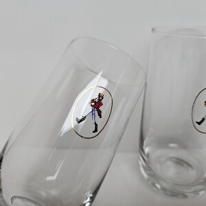 May include: Two clear, tall drinking glasses. Each glass features a gold oval sticker with a figure in a red coat, black boots, and a yellow hat. The glasses are set against a white background.