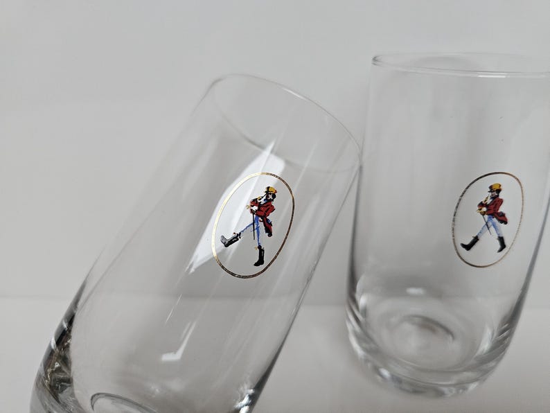 May include: Two clear, tall drinking glasses. Each glass features a gold oval sticker with a figure in a red coat, black boots, and a yellow hat. The glasses are set against a white background.