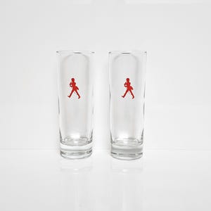 Peut inclure: Deux verres &agrave; boire clairs et hauts, avec une silhouette rouge d'une personne marchant. Les verres sont cylindriques avec un dessus l&eacute;g&egrave;rement &eacute;vas&eacute; et reposent sur une surface blanche.