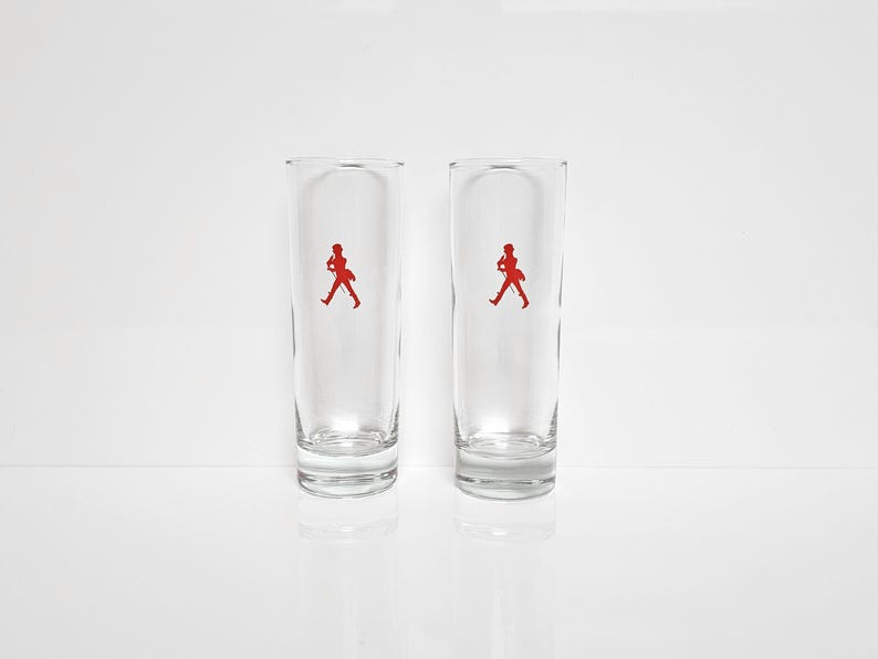 Peut inclure: Deux verres &agrave; boire clairs et hauts, avec une silhouette rouge d'une personne marchant. Les verres sont cylindriques avec un dessus l&eacute;g&egrave;rement &eacute;vas&eacute; et reposent sur une surface blanche.