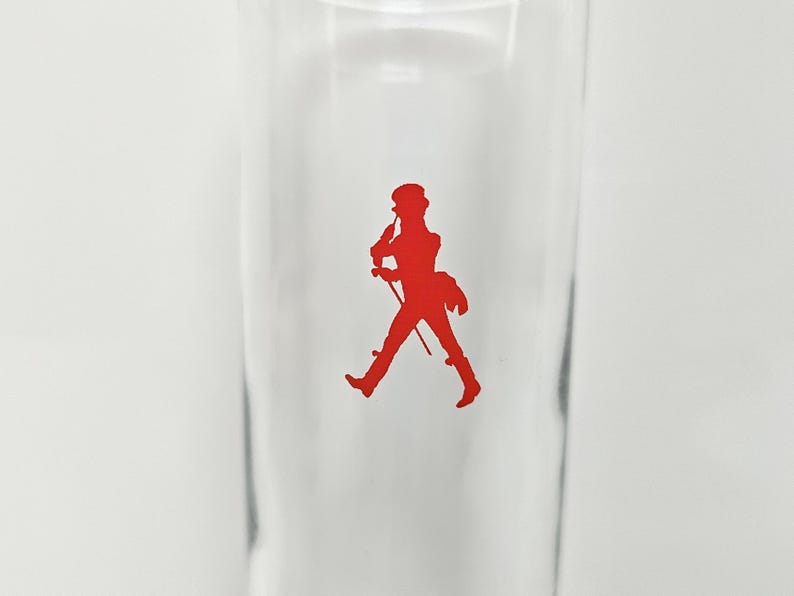 Peut inclure: Un verre transparent avec une silhouette rouge d'un homme marchant, tenant une canne et portant un chapeau. L'image est un gros plan du verre, avec la silhouette centr&eacute;e. Le verre est vide.