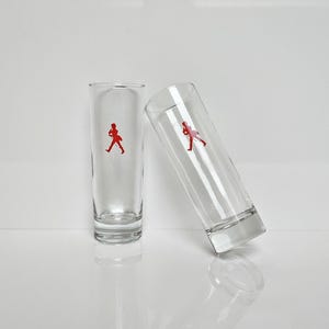 Peut inclure: Deux verres &agrave; shot transparents avec un logo de personnage rouge. Un verre est droit, l'autre est inclin&eacute; contre lui. Les verres sont en verre transparent et ont un design simple et moderne.
