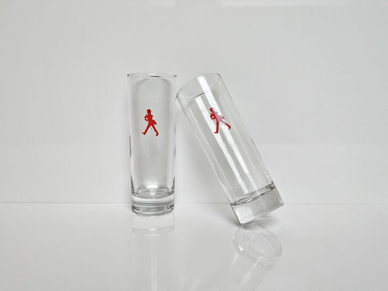 Peut inclure: Deux verres &agrave; shot transparents avec un logo de personnage rouge. Un verre est droit, l'autre est inclin&eacute; contre lui. Les verres sont en verre transparent et ont un design simple et moderne.