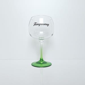 May include: A clear, stemmed gin glass with a green base and stem that fades to clear. The word "Tanqueray" is printed in black script on the glass. The glass is designed for cocktails.