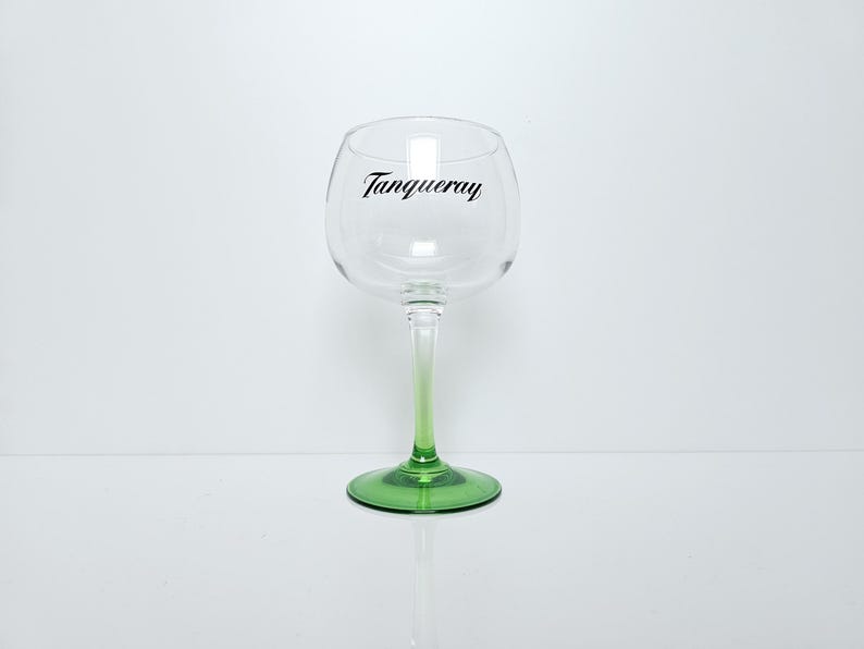 May include: A clear, stemmed gin glass with a green base and stem that fades to clear. The word "Tanqueray" is printed in black script on the glass. The glass is designed for cocktails.