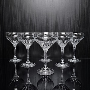 Puede incluir: Un juego de cinco copas de cristal transparente con un cuenco ancho y poco profundo y un tallo corto y delgado. Cada copa presenta un patrón de corte distintivo que irradia desde la base del cuenco, creando un aspecto facetado brillante.