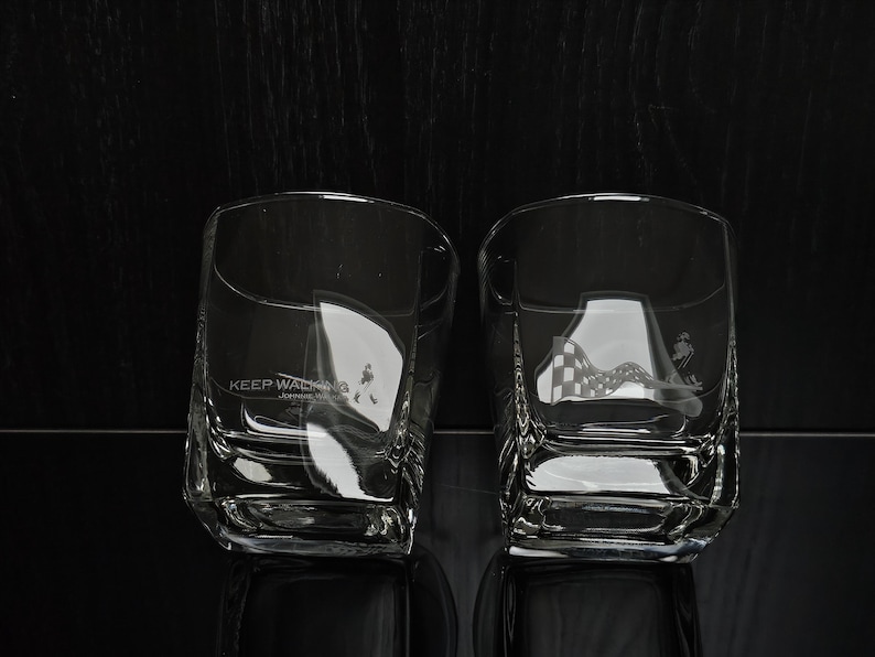 May include: Two clear, square-shaped whiskey glasses. One glass features the text "KEEP WALKING" with a figure walking. The other glass has a checkered flag design and a figure. Both glasses are on a reflective black surface.